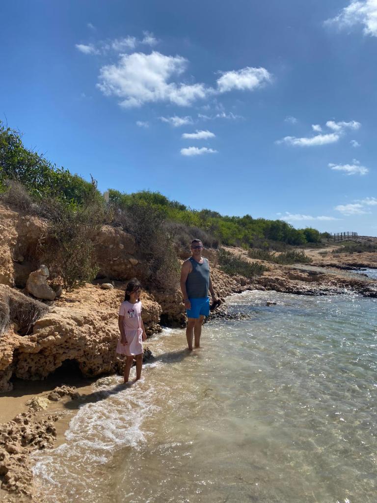Adult and child standing in shallow clear sea water along a rocky Cyprus shoreline