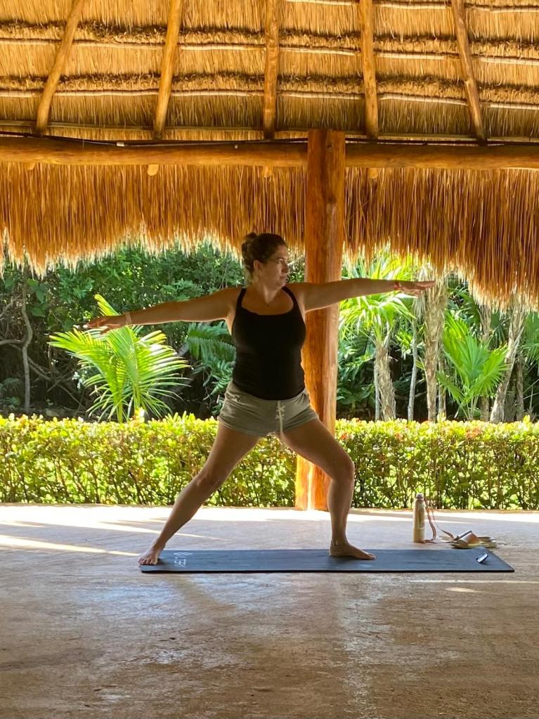 Person practising yoga on a mat under a thatched roof surrounded by greenery in Mexico