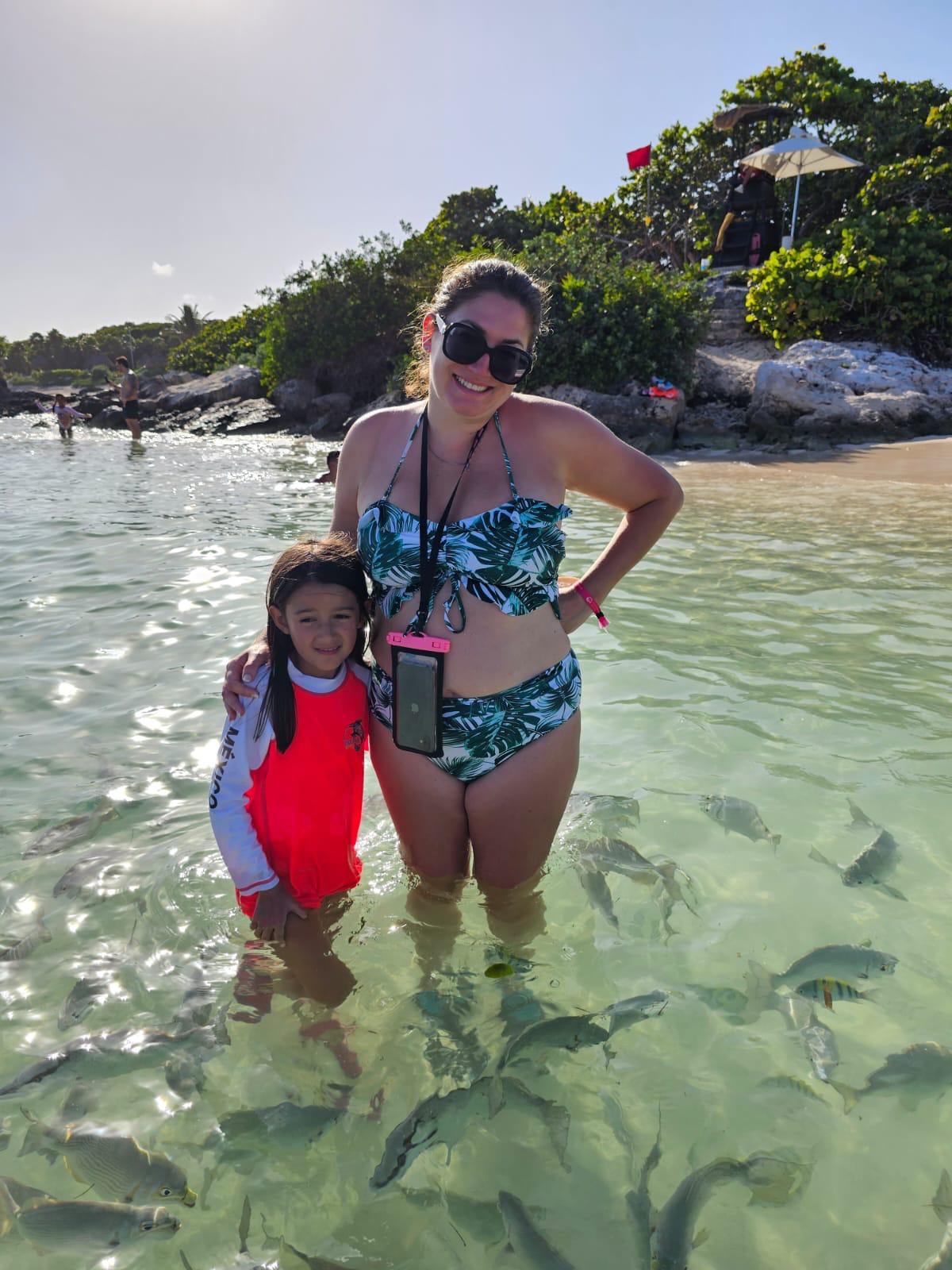 Adult and child standing in shallow clear sea water with fish swimming around them in Mexico