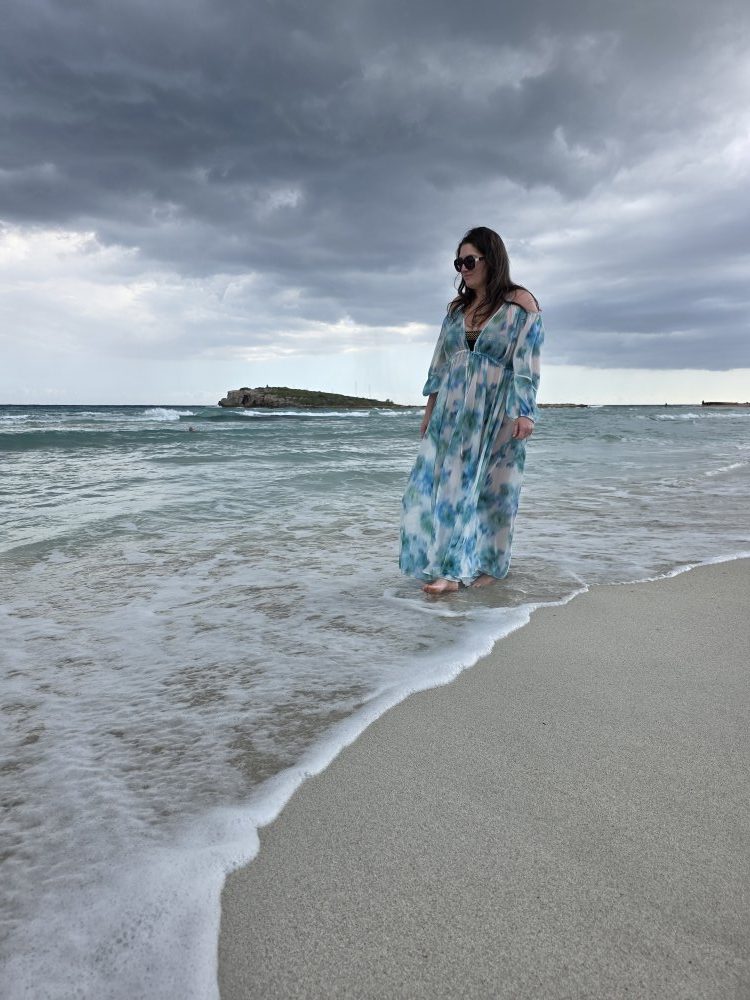 Jodie Steele standing at the edge of the sea in a flowing blue patterned dress beneath a dramatic cloudy sky.